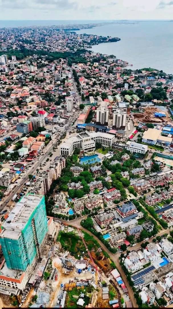 Vertical aerial overview of coastal Conakry cityscape showing modern developments, resort pools, and the ocean shoreline.